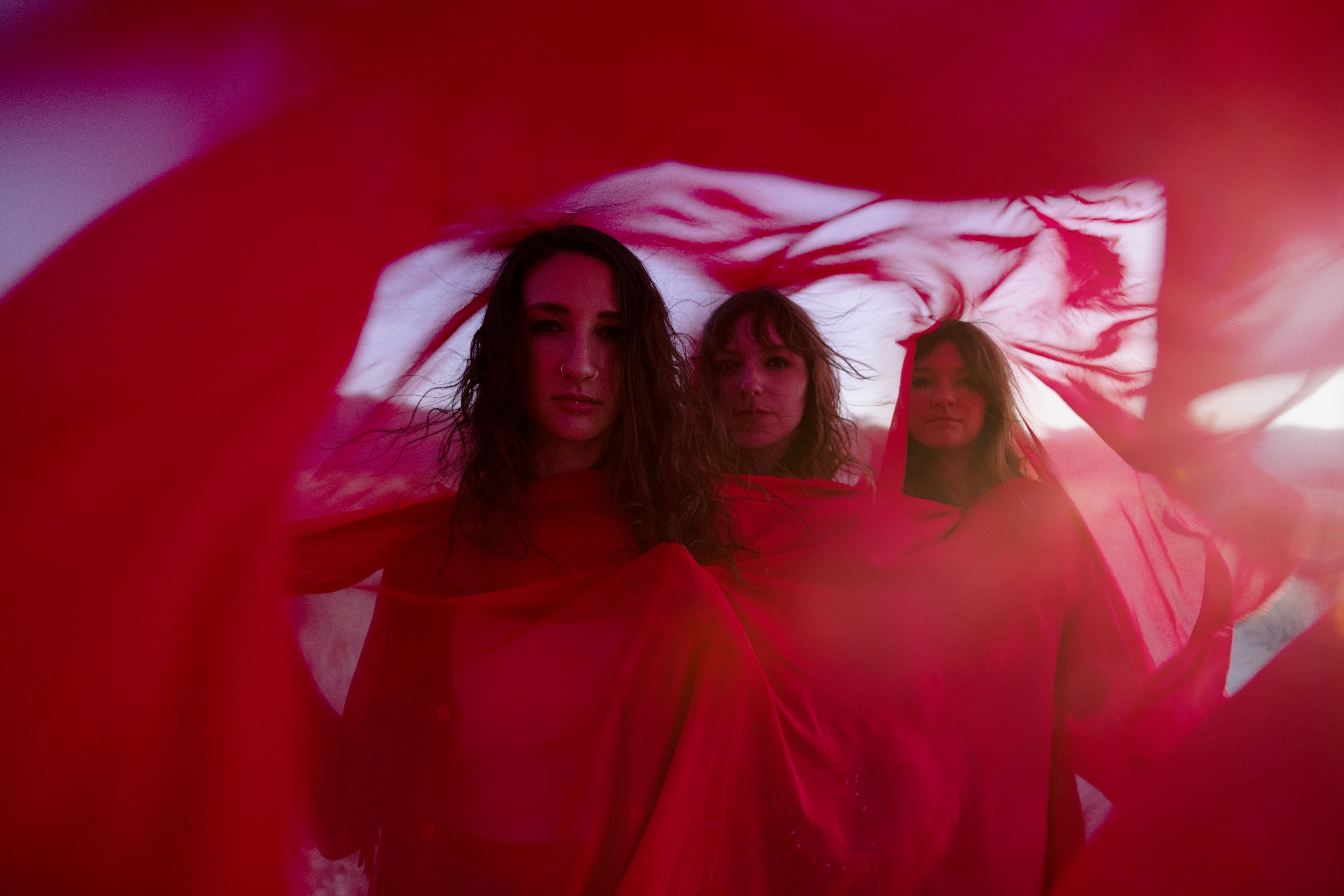 Three women with long, dark hair look directly at the camera, framed by layers of flowing, translucent red fabric that creates a soft, ethereal border. The lighting is warm and hazy, giving the outdoor desert setting a dreamlike, cinematic quality.