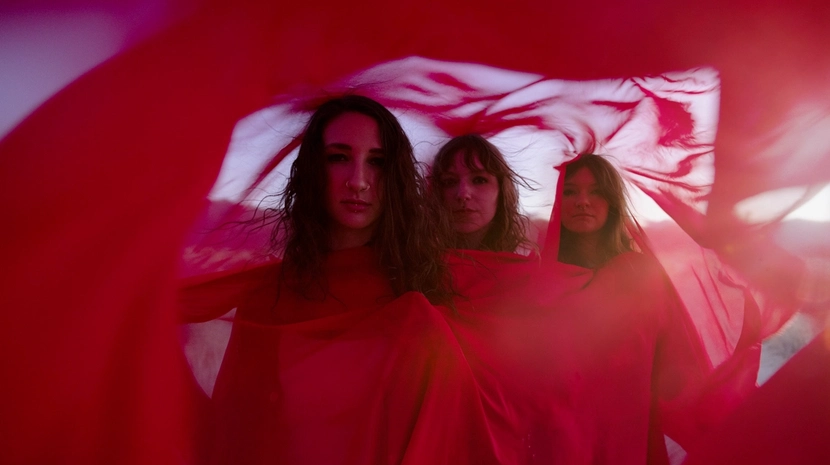 Three women with long, dark hair look directly at the camera, framed by layers of flowing, translucent red fabric that creates a soft, ethereal border. The lighting is warm and hazy, giving the outdoor desert setting a dreamlike, cinematic quality.