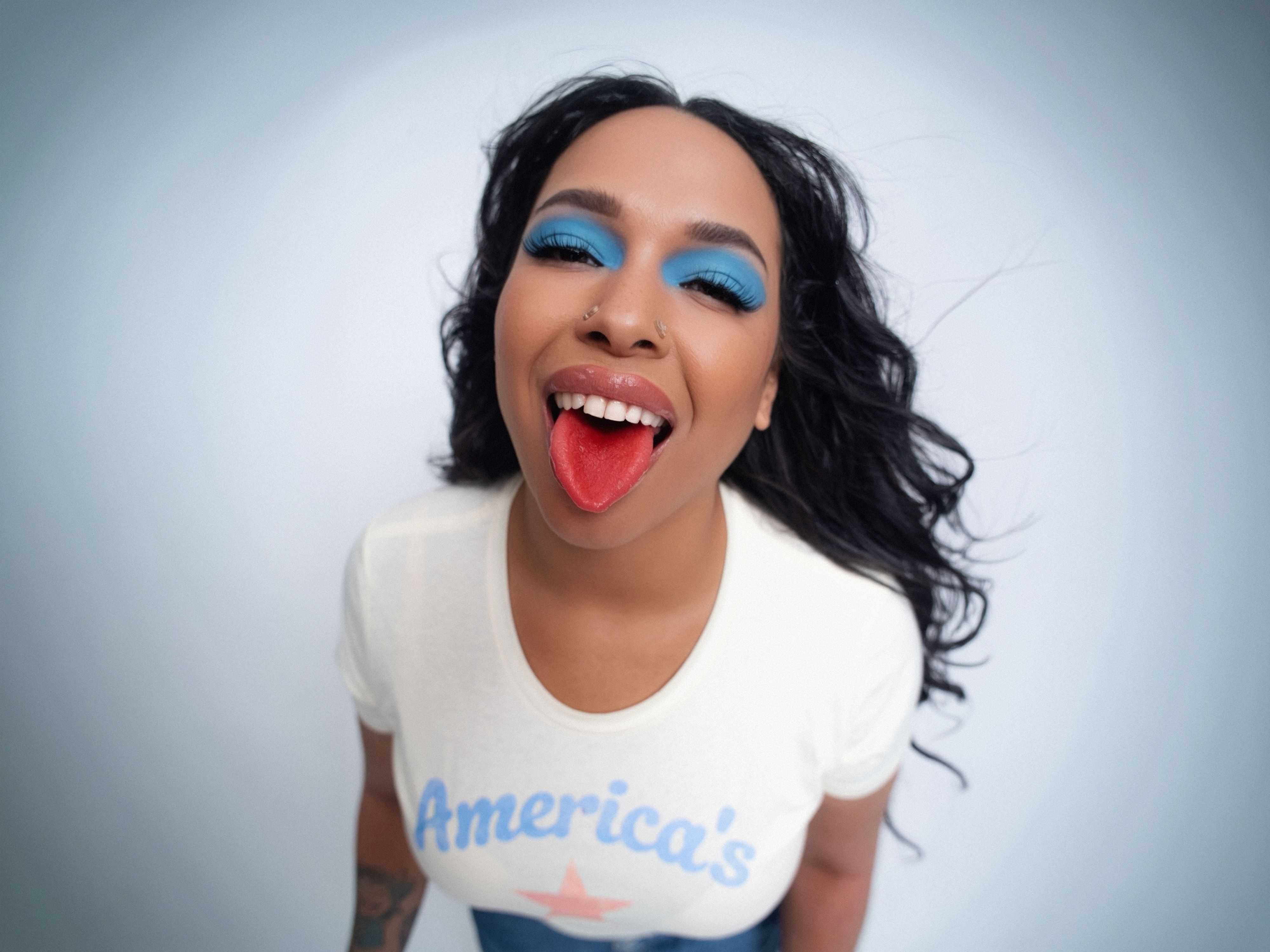 Gemini said A person with long, dark wavy hair and bright blue eyeshadow smiles playfully at a high-angle camera while sticking out a red-stained tongue. They are wearing a white t-shirt with "America's" printed in blue text above a red star, set against a clean, neutral background.