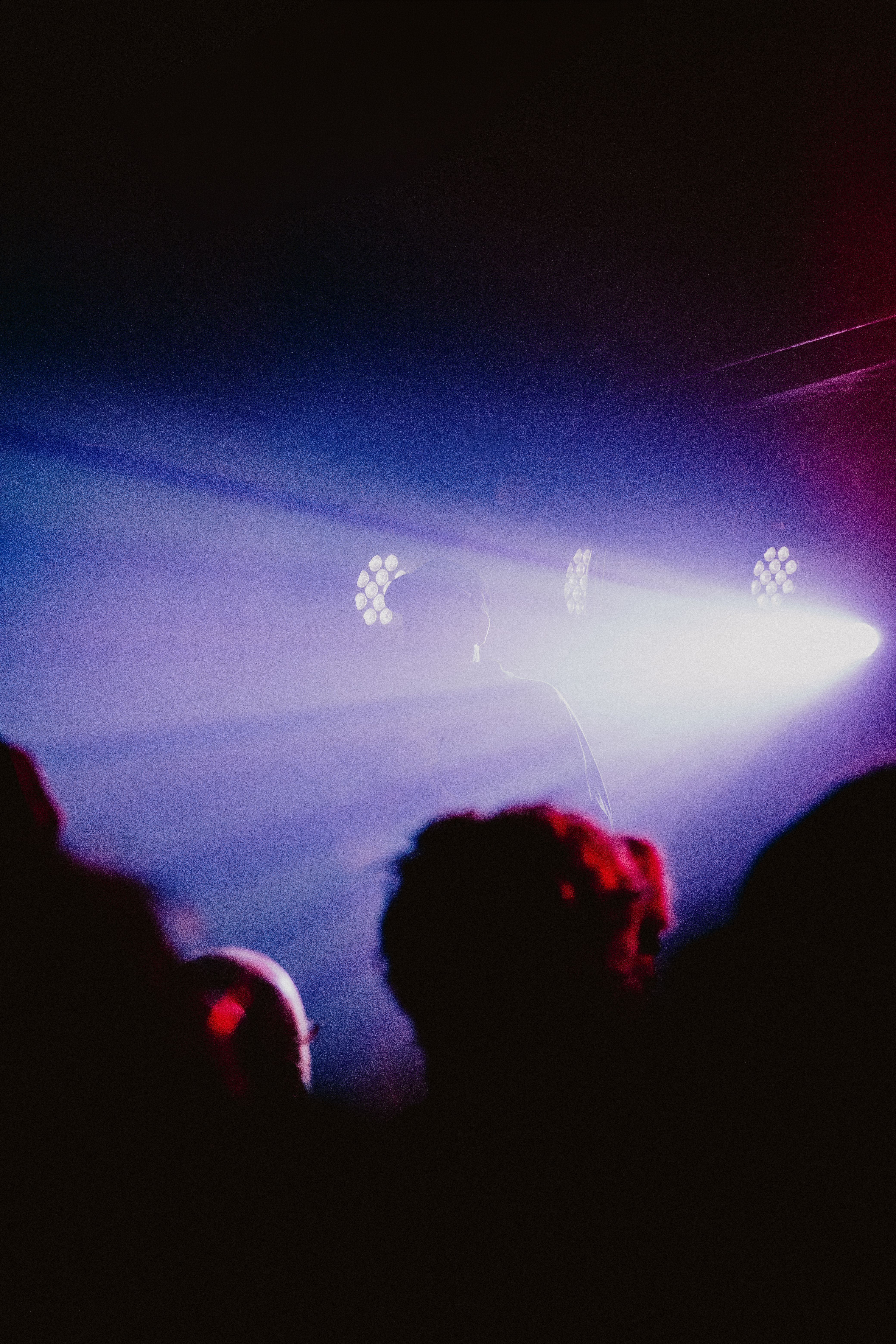 A person is silhouetted against a hazy, deep purple and blue light stage while performing for a crowd. Bright spotlights cut through the fog, creating a moody and atmospheric concert setting.