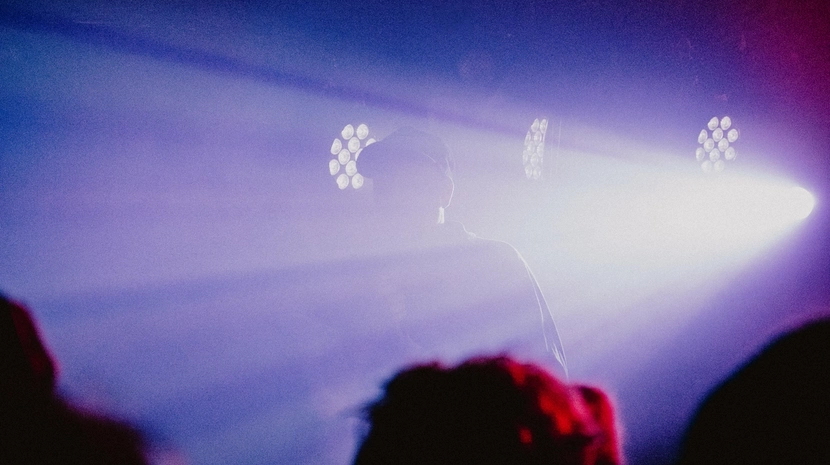 A person is silhouetted against a hazy, deep purple and blue light stage while performing for a crowd. Bright spotlights cut through the fog, creating a moody and atmospheric concert setting.
