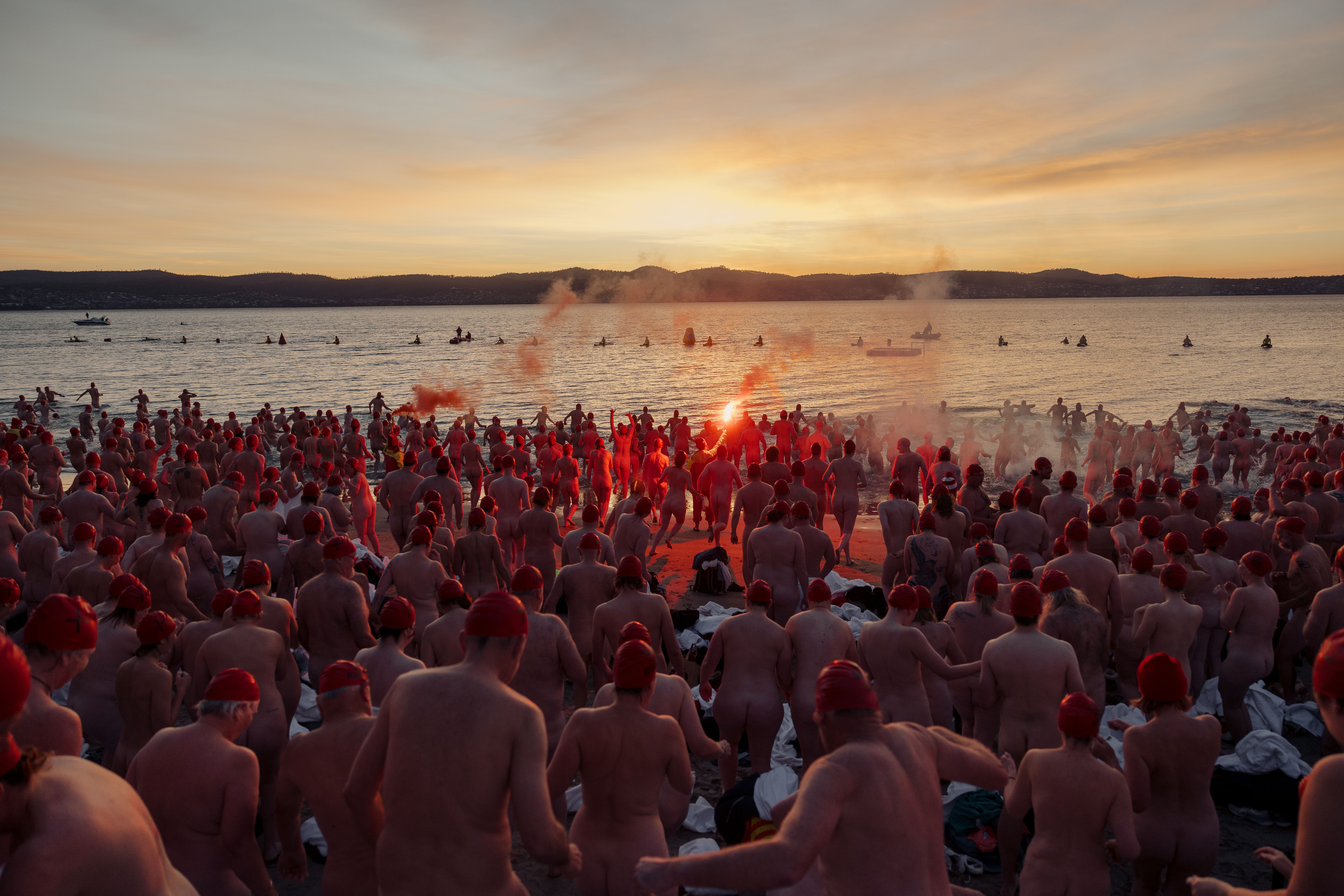 A massive crowd of nude swimmers wearing red caps charges into the ocean at sunrise, led by a person holding a glowing orange flare trailing smoke.