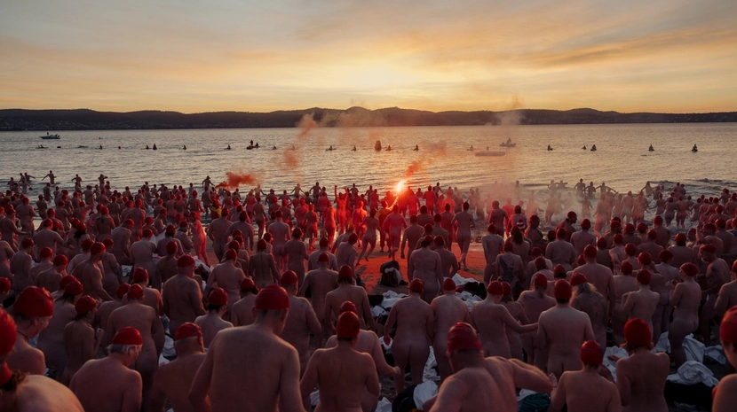 A massive crowd of nude swimmers wearing red caps charges into the ocean at sunrise, led by a person holding a glowing orange flare trailing smoke.