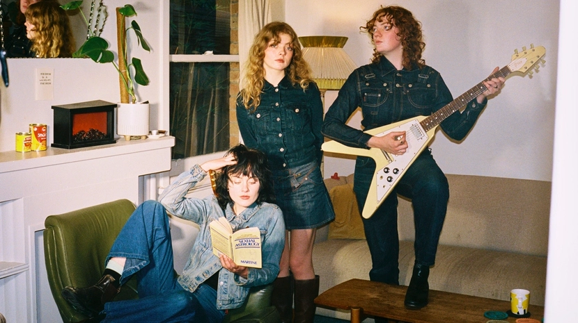 Three people in denim outfits pose in a living room; one sits reading a book, while another stands holding a cream-colored Flying V guitar. The aesthetic is domestic and indie, captured with a film-like quality.