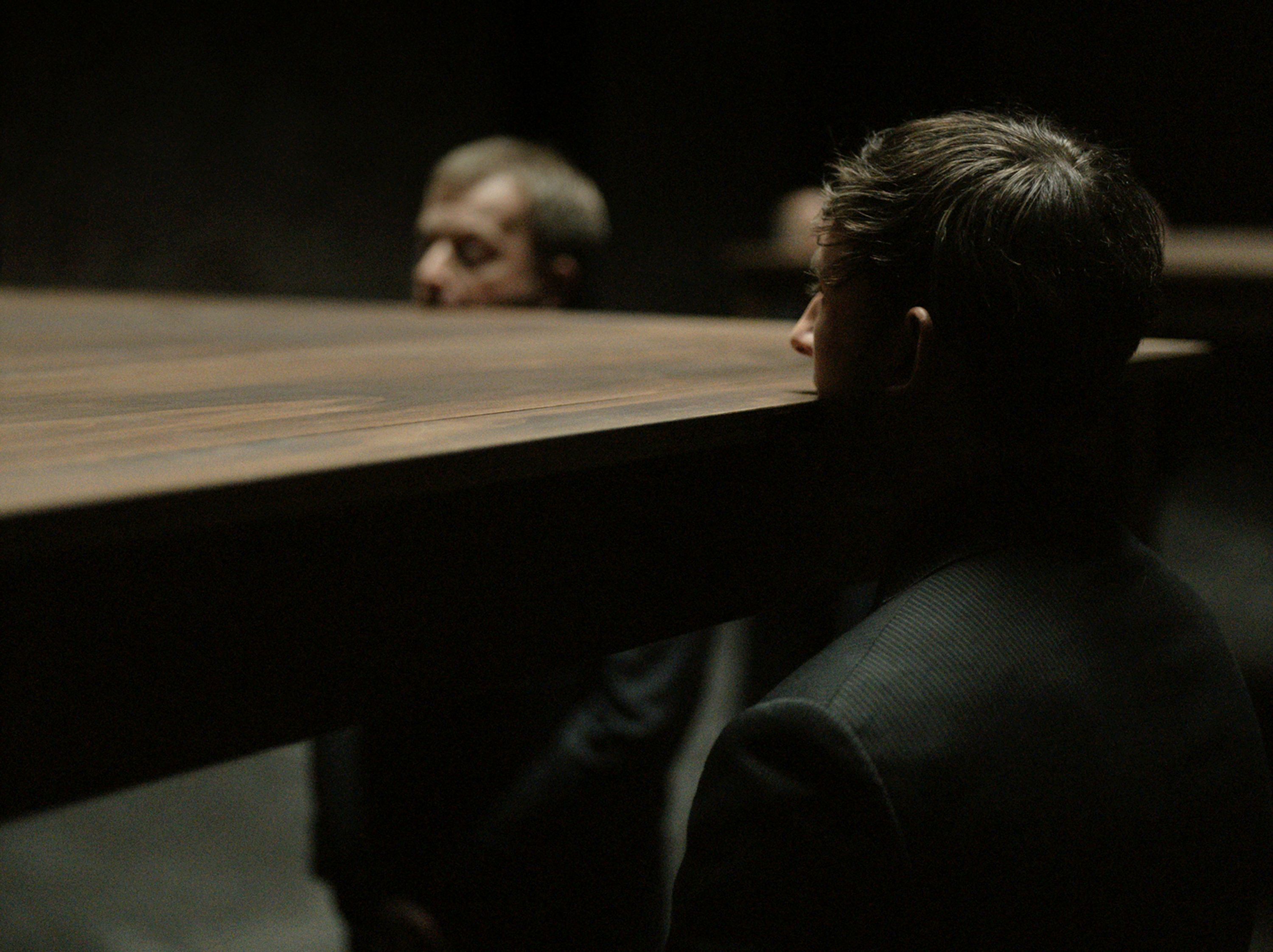 Two men in dark suits are partially obscured by a large, flat wooden table that appears to cut through them at neck level. The lighting is dim, focusing on the surreal and unsettling intersection of the furniture and the human figures.