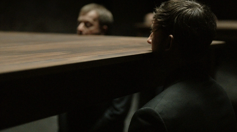 Two men in dark suits are partially obscured by a large, flat wooden table that appears to cut through them at neck level. The lighting is dim, focusing on the surreal and unsettling intersection of the furniture and the human figures.