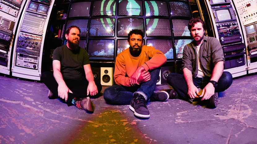 Three men sit on the floor in front of a massive wall of vintage cathode-ray tube televisions displaying a pixelated green graphic. The room is packed with stacks of electronic equipment and rack-mounted gear.