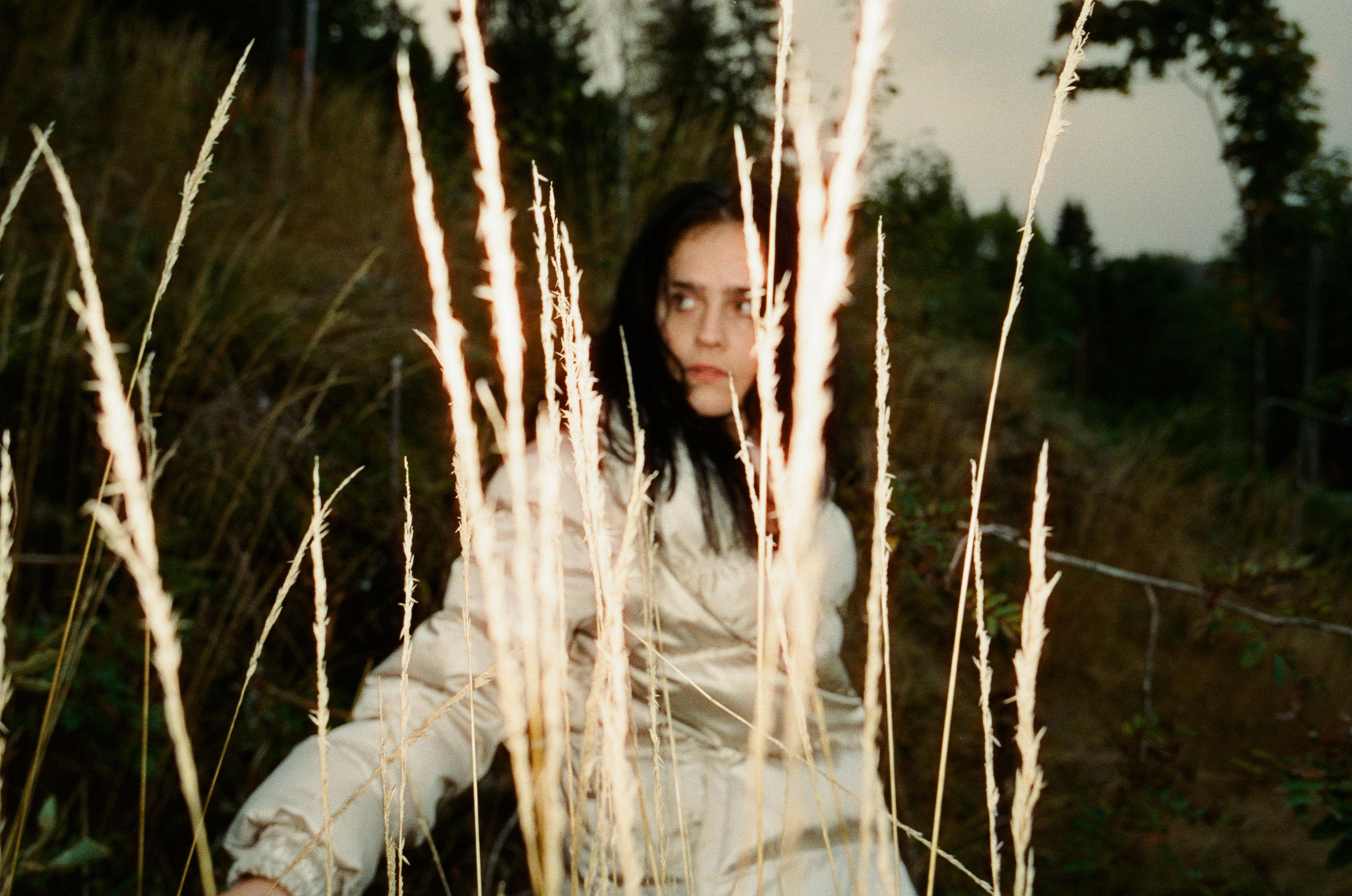 A young woman with dark hair peers cautiously through tall, dry grass in a field at dusk. She wears a light-colored puffer jacket, and the soft, natural lighting creates a cinematic and mysterious mood.