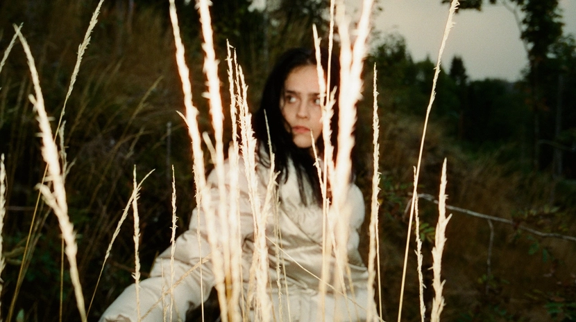 A young woman with dark hair peers cautiously through tall, dry grass in a field at dusk. She wears a light-colored puffer jacket, and the soft, natural lighting creates a cinematic and mysterious mood.