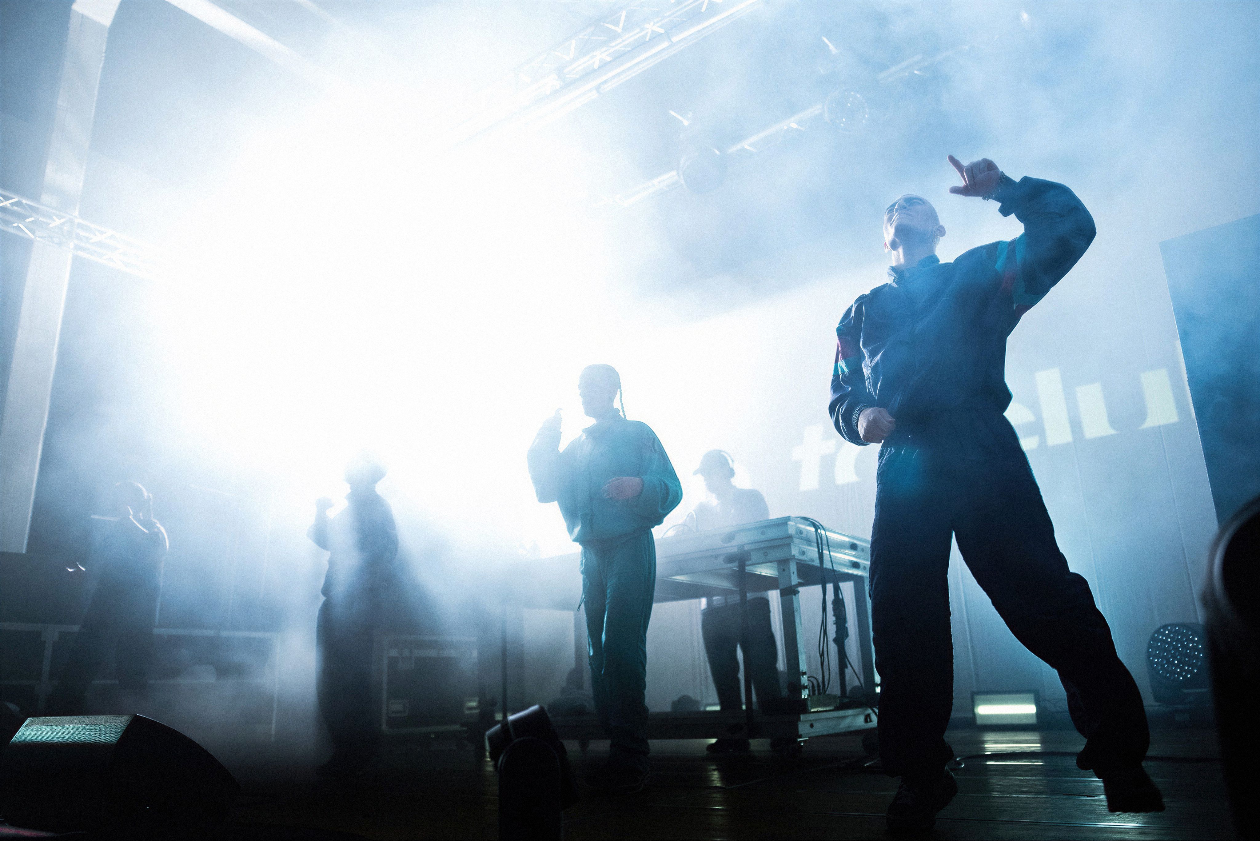 Silhouette figures of performers in tracksuits dance on a hazy, backlit stage filled with bright white light and theatrical smoke. A DJ stands behind a table in the background, while the energetic atmosphere captures the essence of a gabber or hardcore techno event.