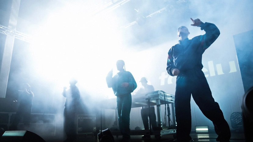 Silhouette figures of performers in tracksuits dance on a hazy, backlit stage filled with bright white light and theatrical smoke. A DJ stands behind a table in the background, while the energetic atmosphere captures the essence of a gabber or hardcore techno event.