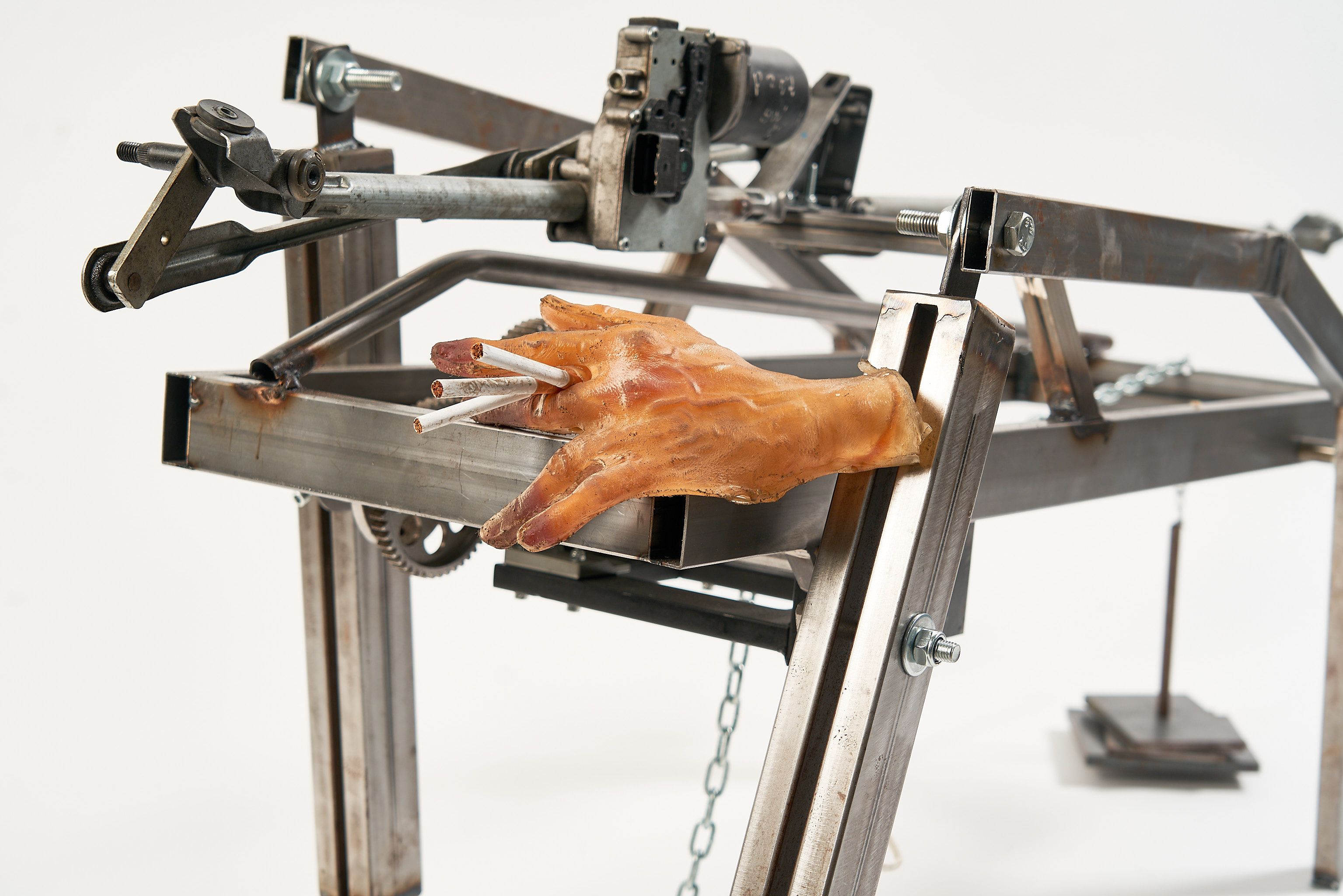 A kinetic metal sculpture made of industrial beams and gears, featuring a realistic prosthetic hand holding three cigarettes between its fingers. The mechanical frame appears rugged and functional, set against a stark white background.