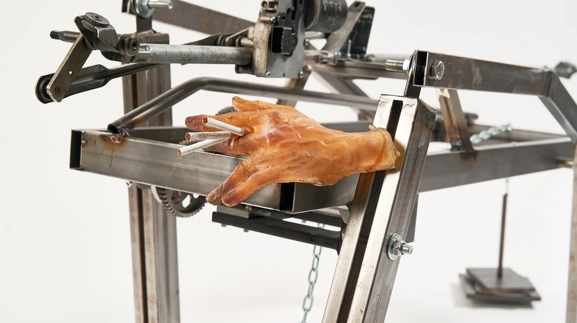 A kinetic metal sculpture made of industrial beams and gears, featuring a realistic prosthetic hand holding three cigarettes between its fingers. The mechanical frame appears rugged and functional, set against a stark white background.