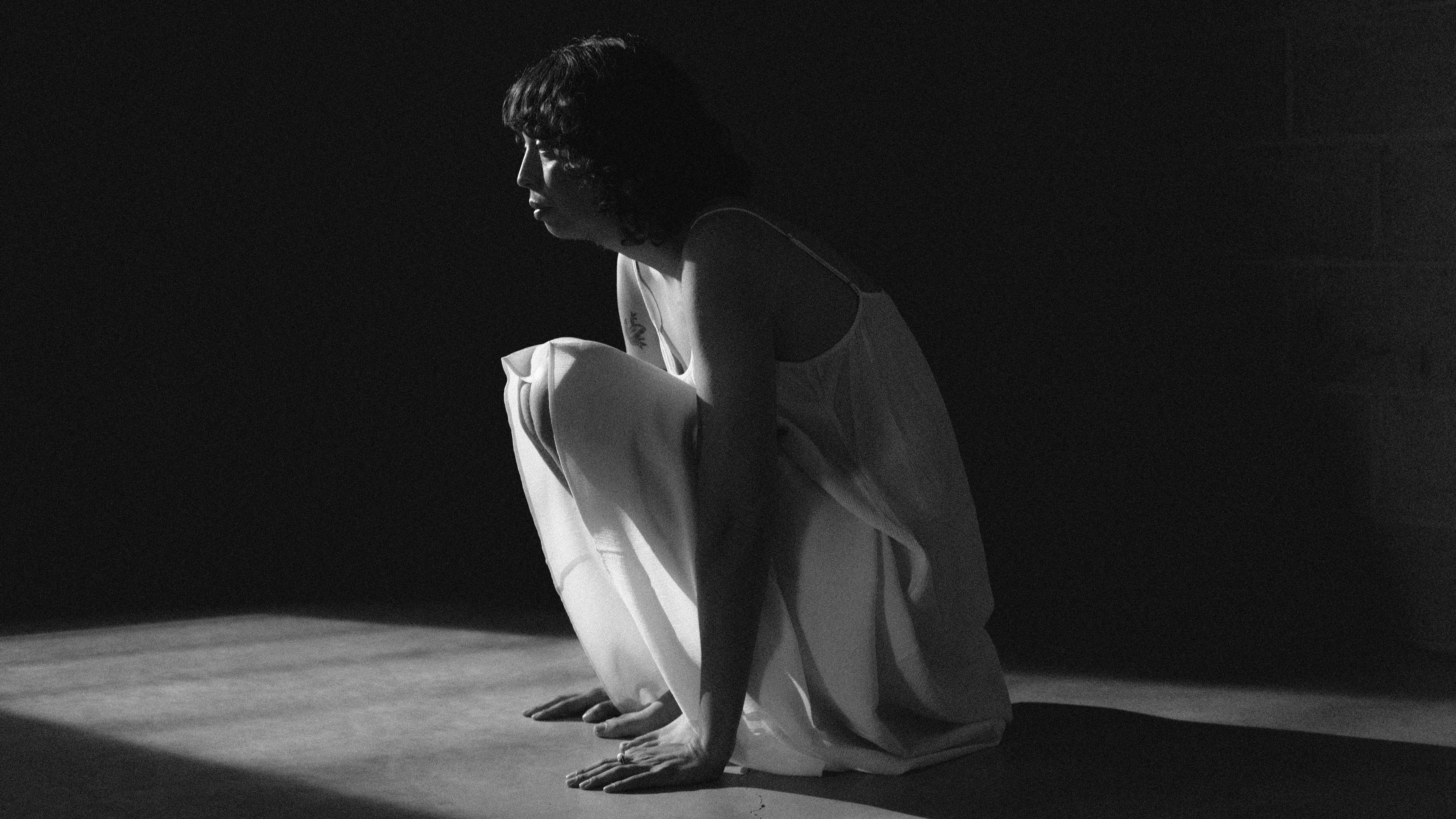 A graining black-and-white photograph shows a person in a light-colored slip dress crouching on a floor. They are positioned in a harsh beam of light that cuts through an otherwise dark, shadowed space.