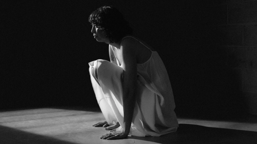 A graining black-and-white photograph shows a person in a light-colored slip dress crouching on a floor. They are positioned in a harsh beam of light that cuts through an otherwise dark, shadowed space.