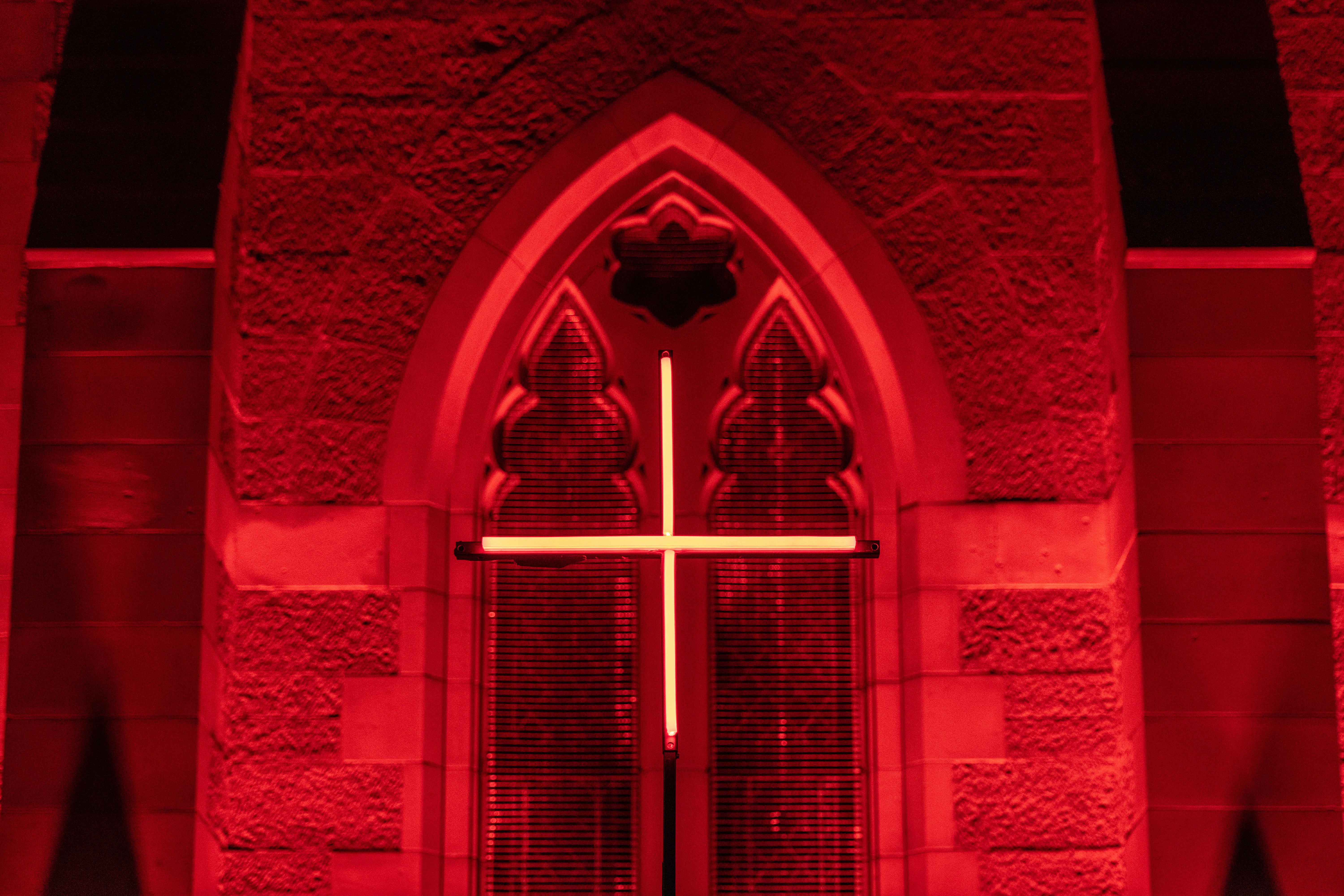 A close-up of a Gothic-style arched window in a stone building, glowing red with a minimalist neon cross centered in the frame.