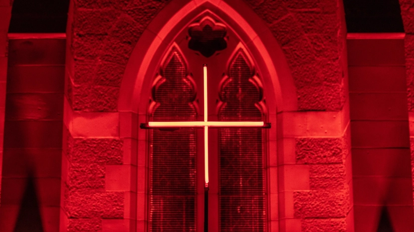 A close-up of a Gothic-style arched window in a stone building, glowing red with a minimalist neon cross centered in the frame.