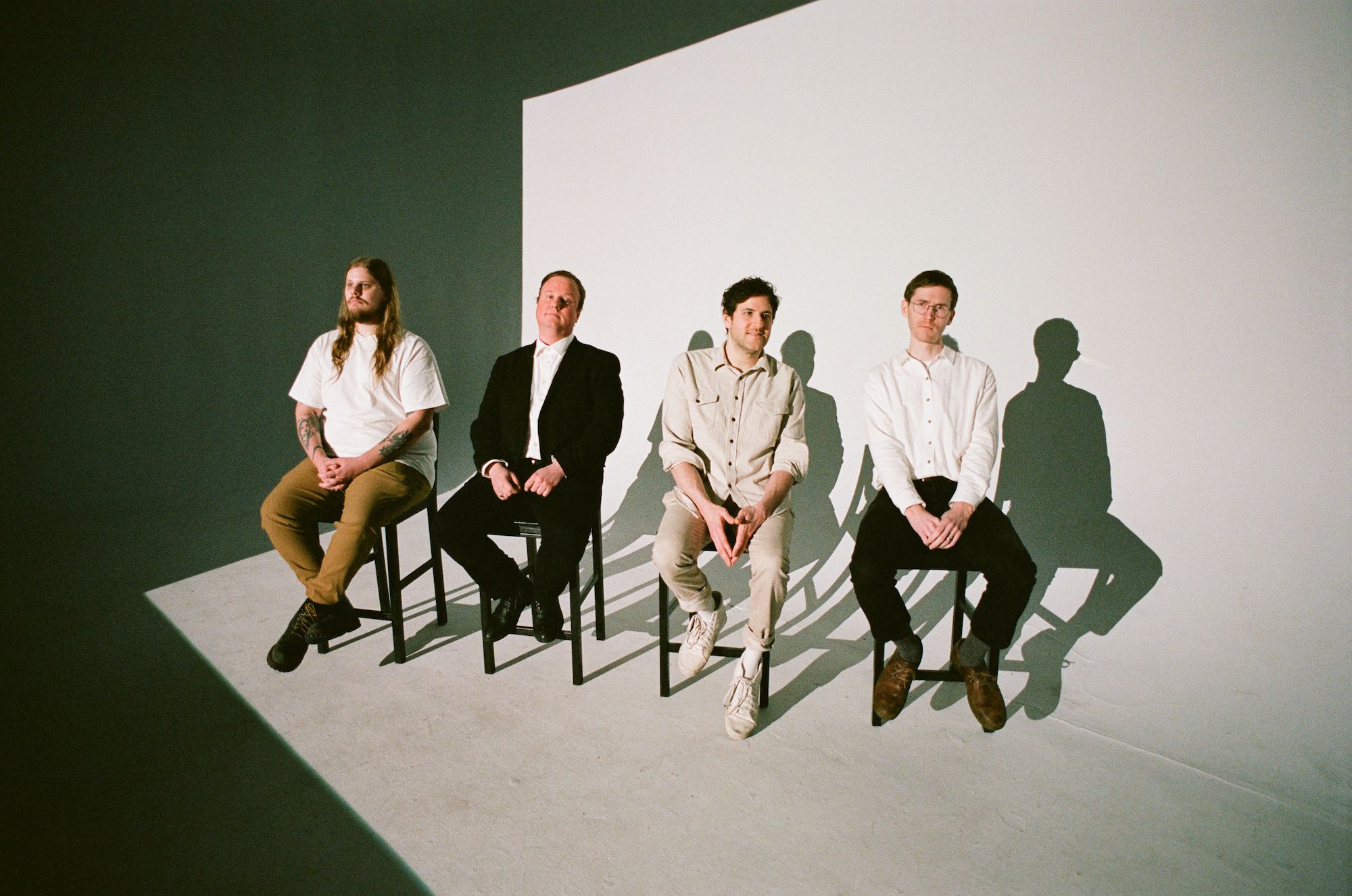 Four men sit in a row on black stools against a stark white backdrop in a studio setting. They are dressed in simple, neutral-toned clothing, and their shadows are cast sharply against the wall behind them.
