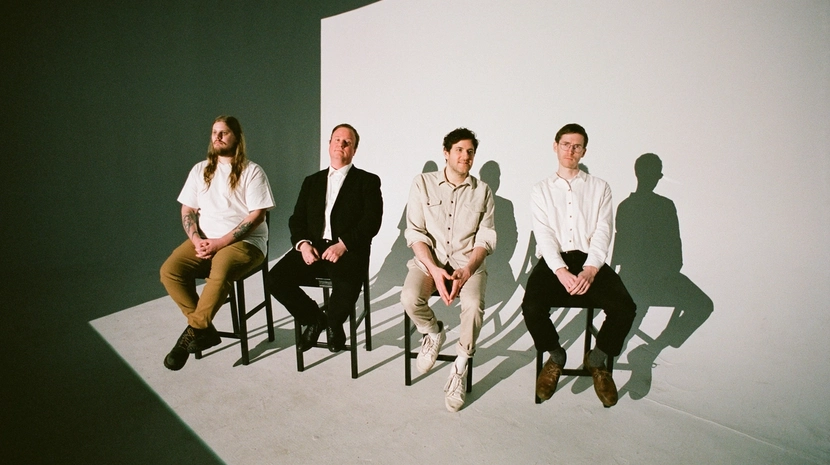 Four men sit in a row on black stools against a stark white backdrop in a studio setting. They are dressed in simple, neutral-toned clothing, and their shadows are cast sharply against the wall behind them.