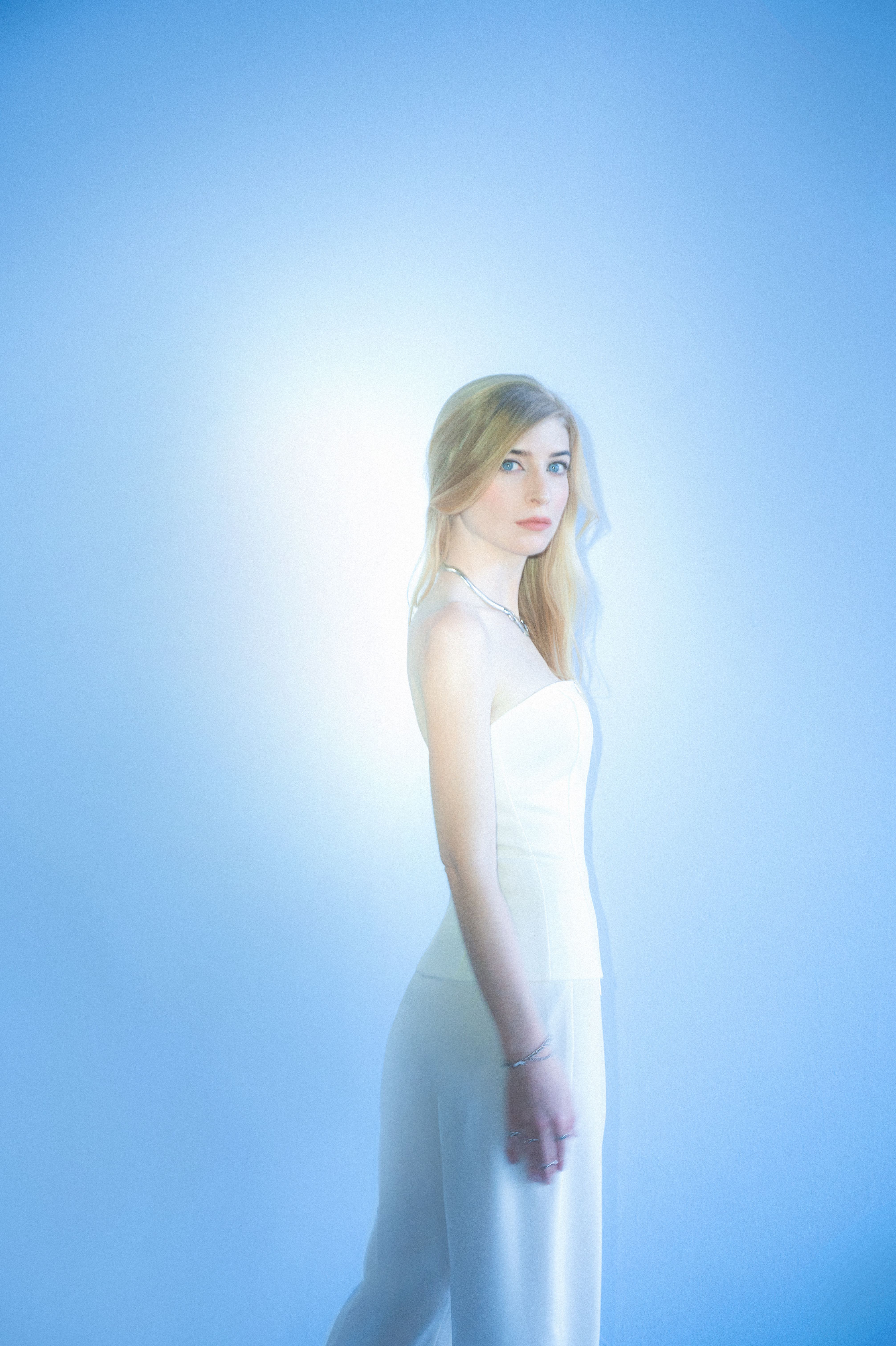 A woman with long blonde hair and blue eyes poses in a white strapless top against a soft, glowing blue background. The lighting is ethereal and bright, creating a halo effect around her silhouette.