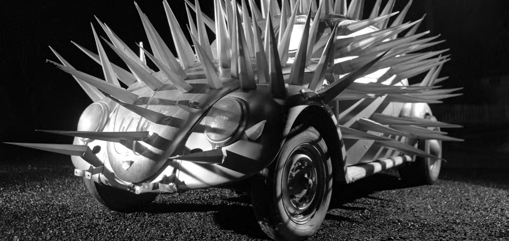 A black-and-white shot of a Volkswagen Beetle completely covered in long, menacing metal spikes. The car sits on a gravel surface at night, appearing like a mechanical porcupine under harsh, dramatic lighting.