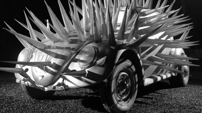 A black-and-white shot of a Volkswagen Beetle completely covered in long, menacing metal spikes. The car sits on a gravel surface at night, appearing like a mechanical porcupine under harsh, dramatic lighting.