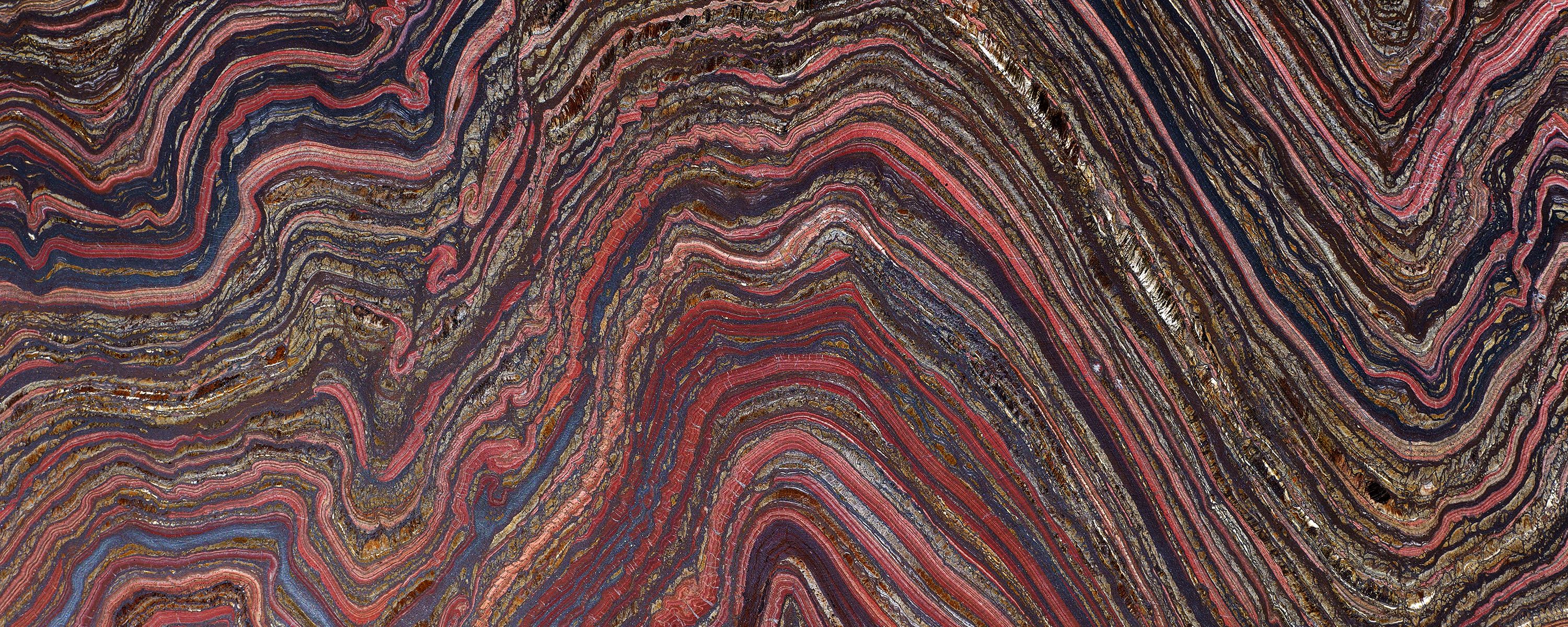 A high-resolution close-up of a rock cross-section showing intricate, wavy bands of deep red, iron-grey, and shimmering gold. The geological layers create a dense, rhythmic pattern of undulating lines across the entire frame.