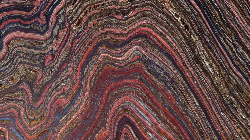 A high-resolution close-up of a rock cross-section showing intricate, wavy bands of deep red, iron-grey, and shimmering gold. The geological layers create a dense, rhythmic pattern of undulating lines across the entire frame.