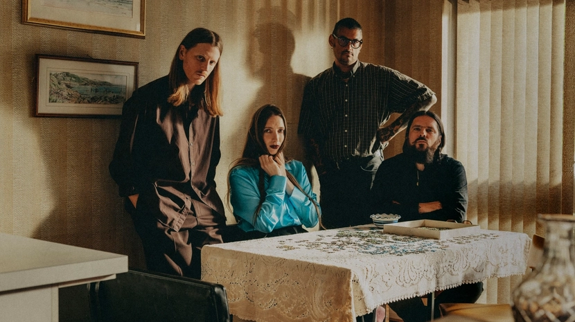 Four band members pose in a vintage-style room with patterned wallpaper and a lace-covered table featuring a jigsaw puzzle. The lighting is soft and warm, giving the scene a retro, cinematic feel.