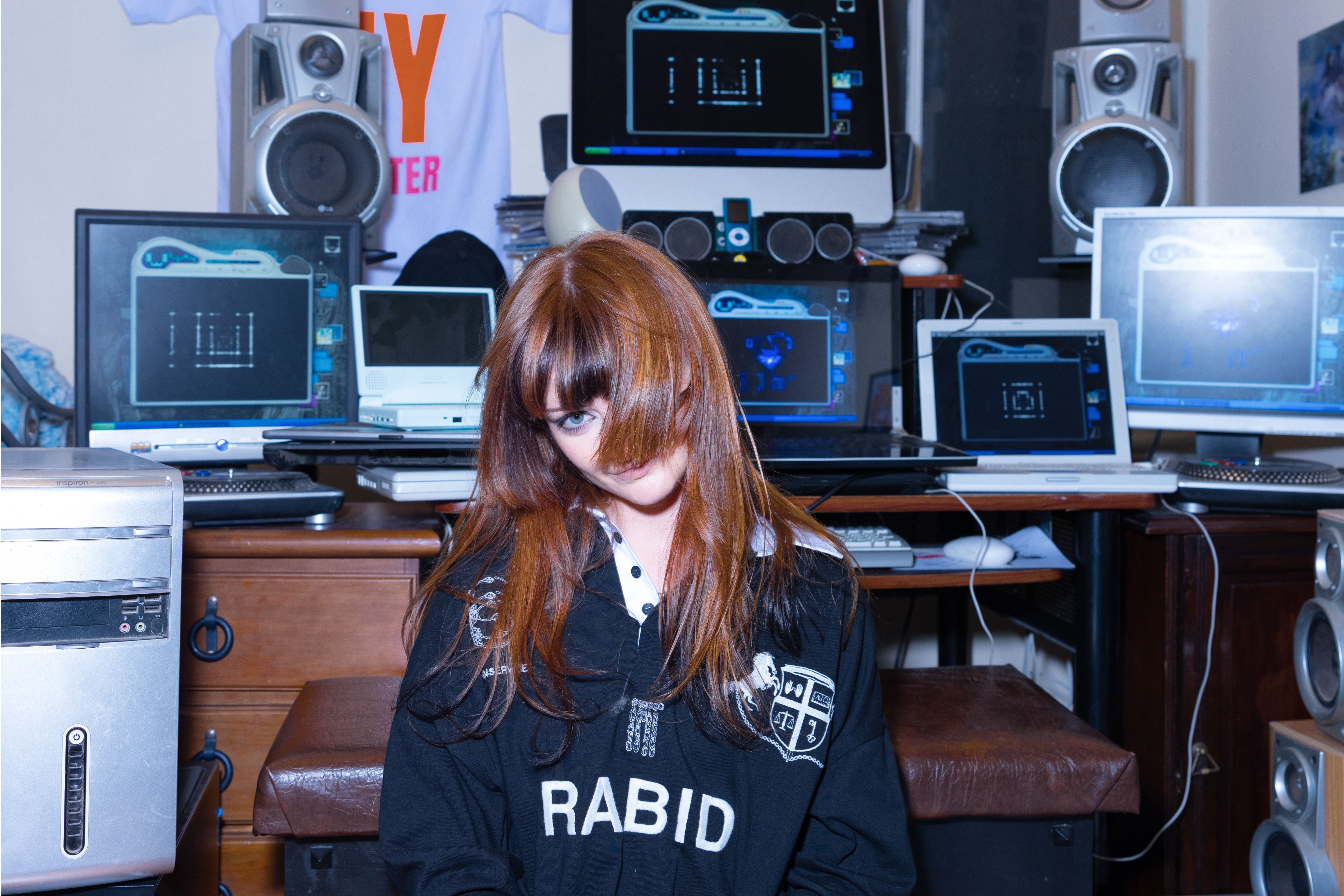 Gemini said A young woman with long reddish-brown hair and bangs sits in a room packed with vintage electronics, including several desktop monitors, laptops, and large speakers. She wears a black sweatshirt with the word "RABID" printed on it, posing amidst the cluttered, Y2K-aesthetic technology setup.
