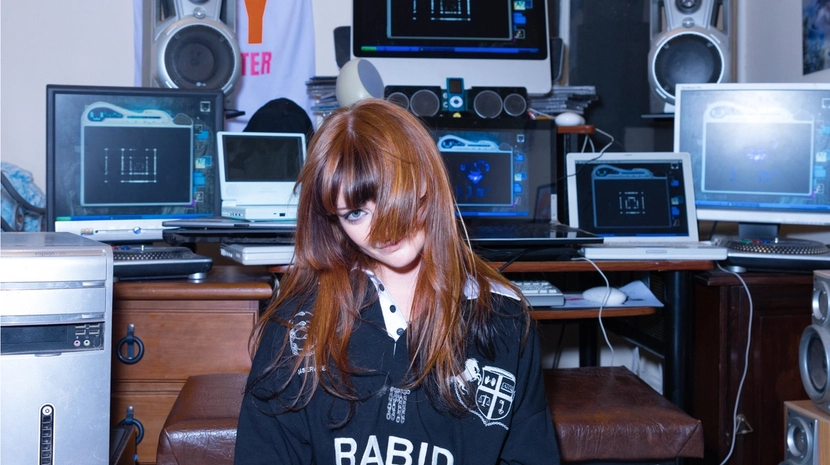 Gemini said A young woman with long reddish-brown hair and bangs sits in a room packed with vintage electronics, including several desktop monitors, laptops, and large speakers. She wears a black sweatshirt with the word "RABID" printed on it, posing amidst the cluttered, Y2K-aesthetic technology setup.