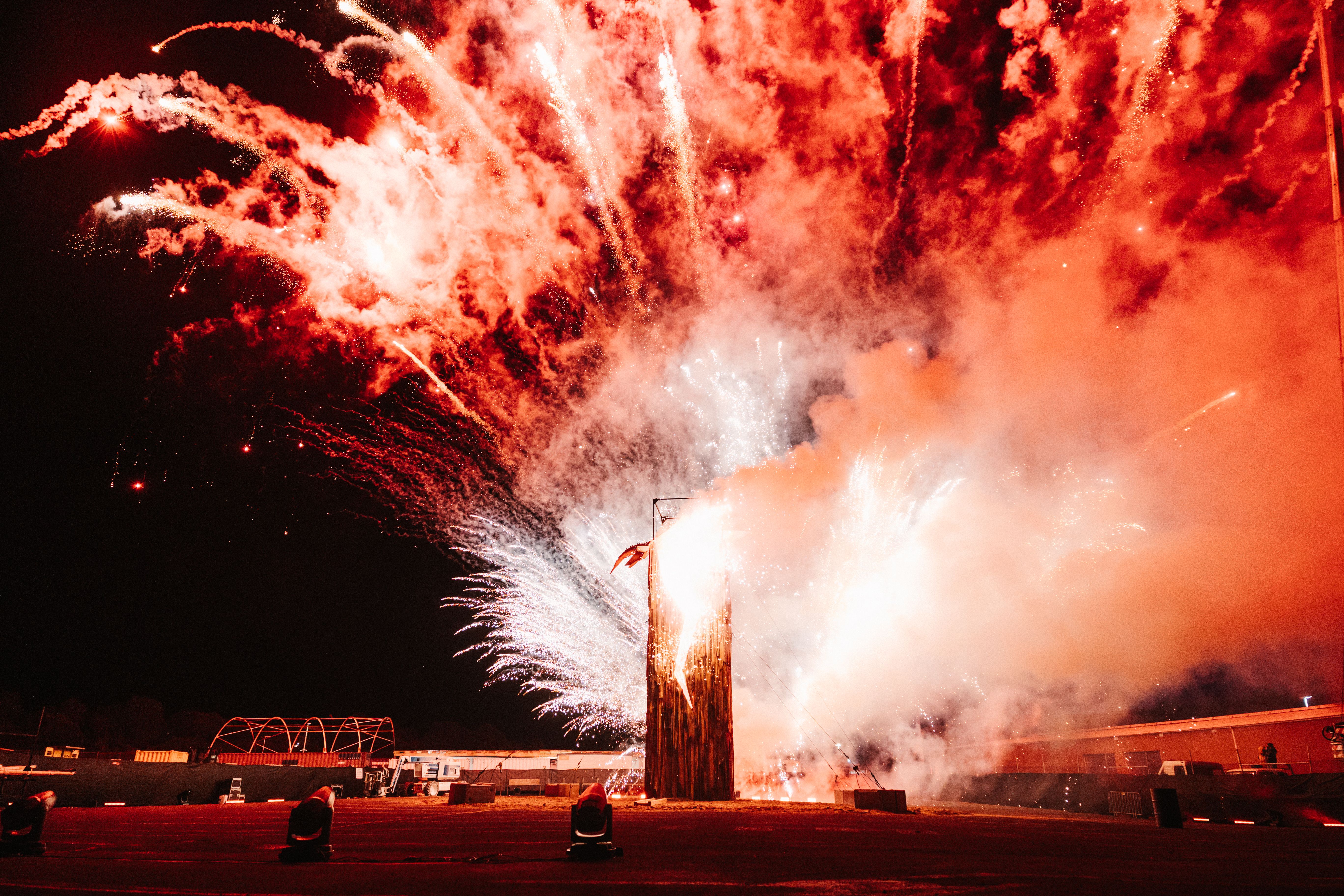 Vibrant white and red fireworks explode behind a tall wooden structure at night, filling the sky with thick, glowing smoke and bright light trails.