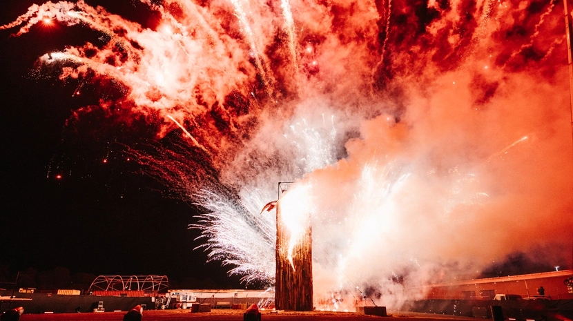 Vibrant white and red fireworks explode behind a tall wooden structure at night, filling the sky with thick, glowing smoke and bright light trails.