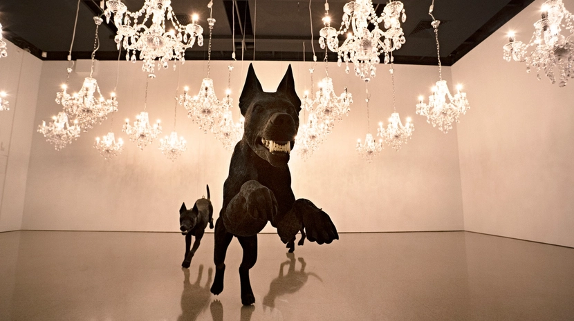Two large, realistic sculptures of black dogs appear to charge forward in a gallery space. The room is dramatically lit by dozens of ornate crystal chandeliers hanging at various heights from the ceiling.
