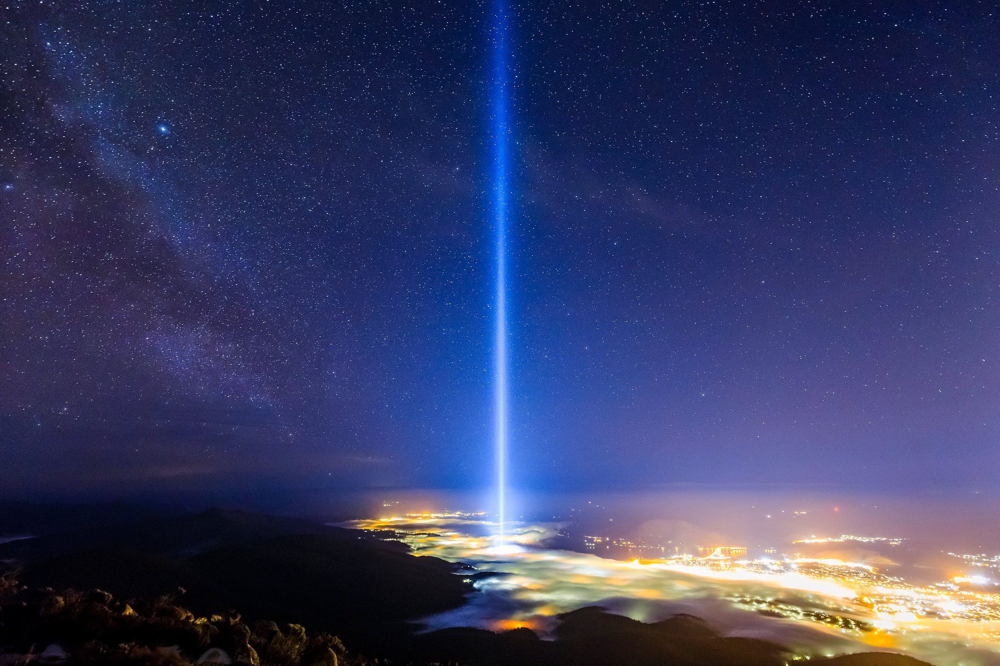 A powerful vertical beam of white light shoots directly into a starry night sky from a glowing city nestled in a valley of clouds. The Milky Way is visible to the left, and the scene is captured from a high, rocky vantage point overlooking the landscape.