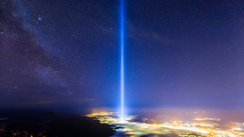 A powerful vertical beam of white light shoots directly into a starry night sky from a glowing city nestled in a valley of clouds. The Milky Way is visible to the left, and the scene is captured from a high, rocky vantage point overlooking the landscape.