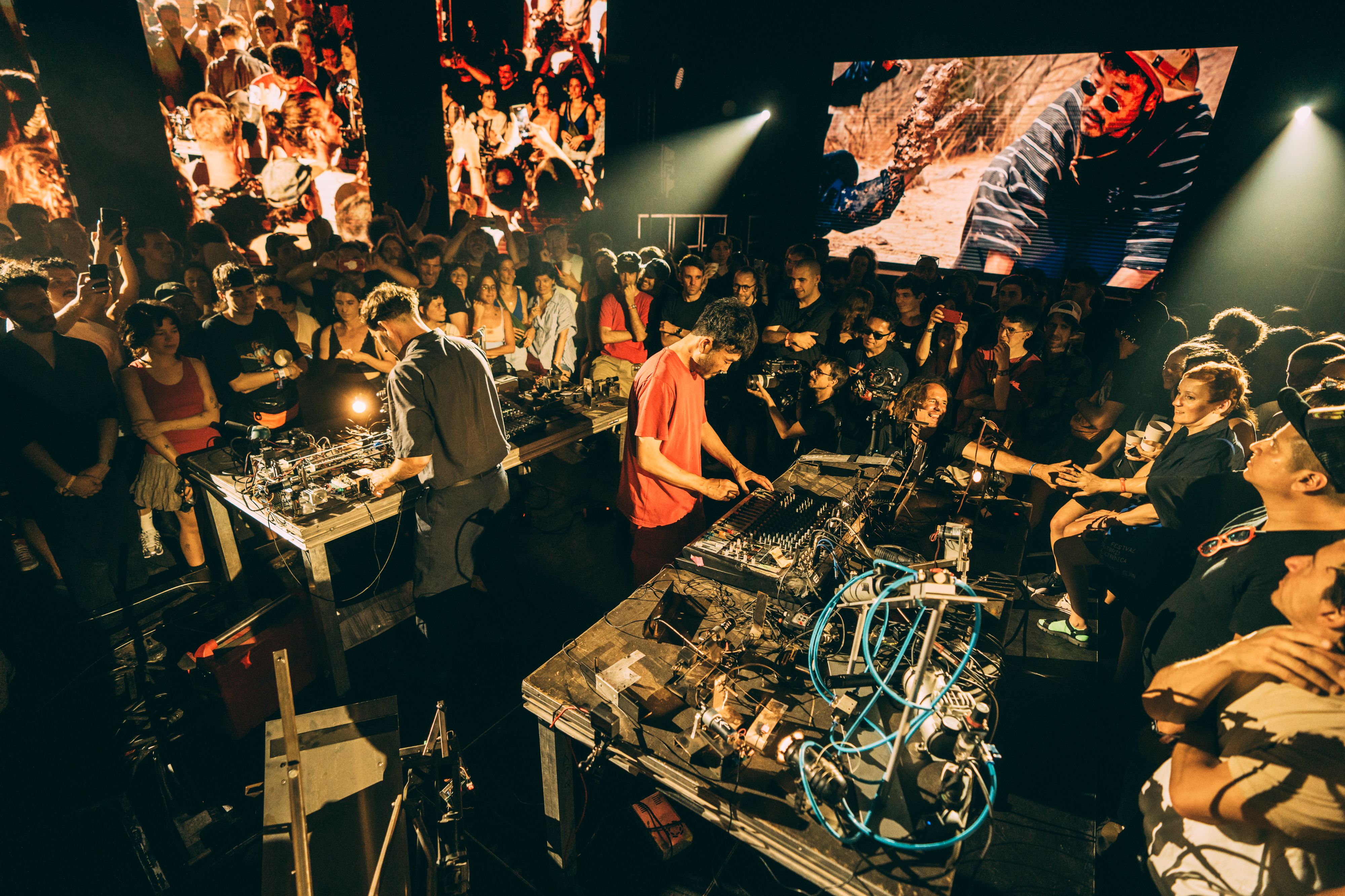 Gemini said Two electronic musicians perform in the center of a crowded, dimly lit venue, surrounded by fans and tables overflowing with synthesizers, wires, and gear. Large screens in the background display vibrant visuals and close-ups of the performance, adding to the high-energy, immersive atmosphere.