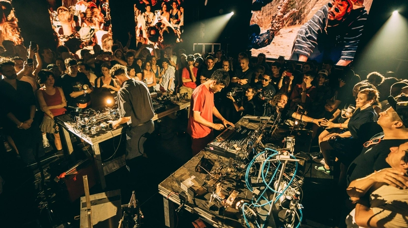 Gemini said Two electronic musicians perform in the center of a crowded, dimly lit venue, surrounded by fans and tables overflowing with synthesizers, wires, and gear. Large screens in the background display vibrant visuals and close-ups of the performance, adding to the high-energy, immersive atmosphere.