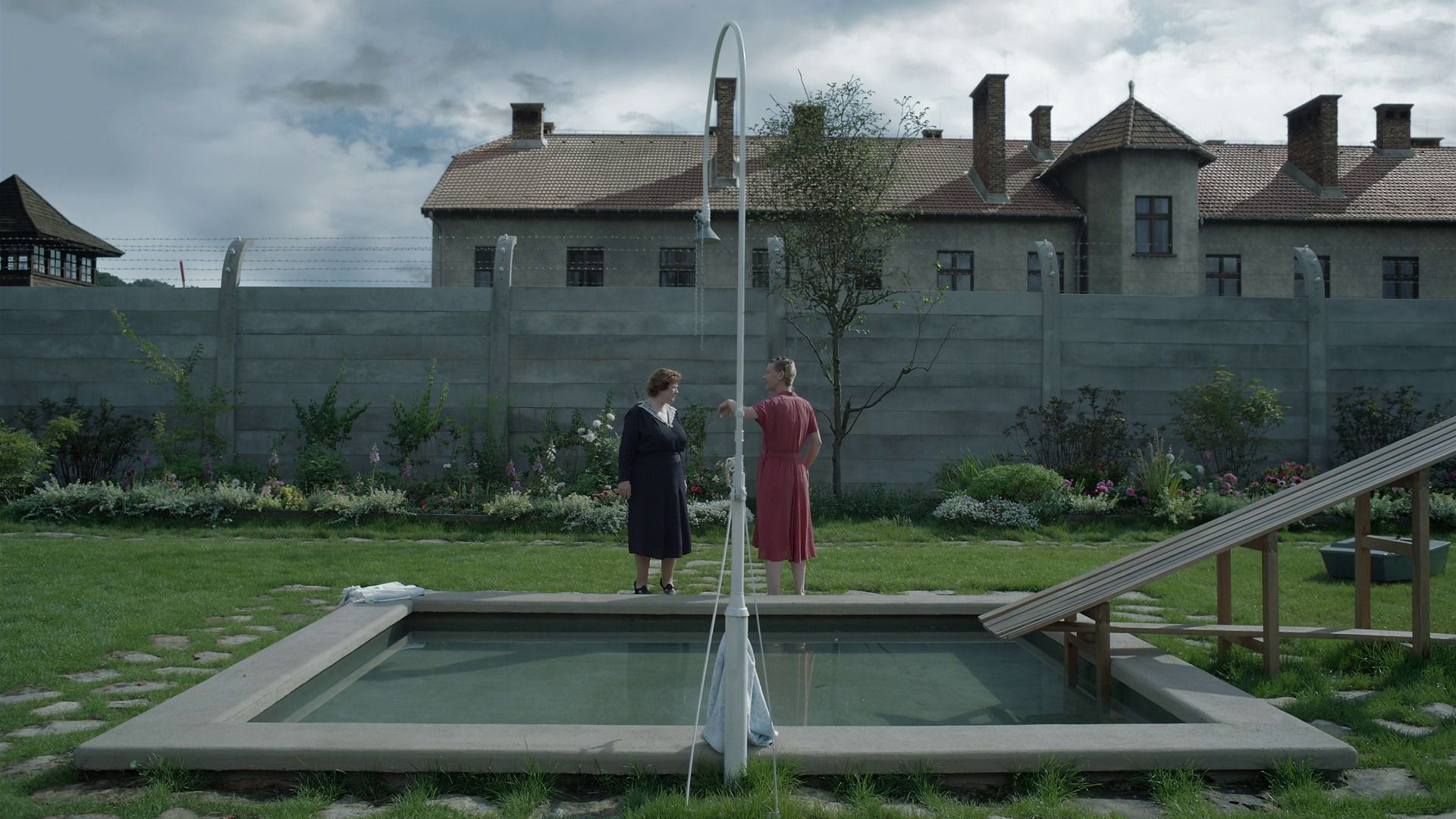 A wide, symmetrical shot of two women talking in a manicured garden next to a small swimming pool and slide. In the background, a high concrete wall topped with barbed wire separates the lush yard from the somber, institutional buildings of a concentration camp.