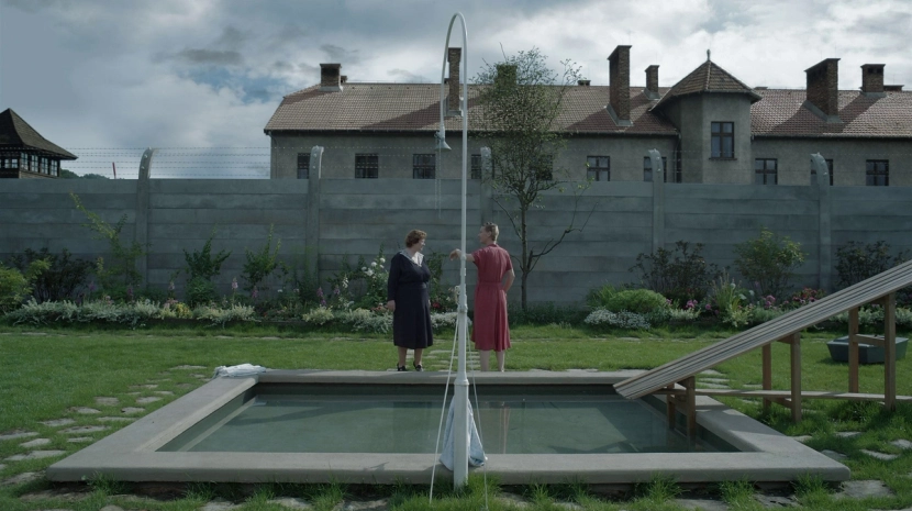 A wide, symmetrical shot of two women talking in a manicured garden next to a small swimming pool and slide. In the background, a high concrete wall topped with barbed wire separates the lush yard from the somber, institutional buildings of a concentration camp.