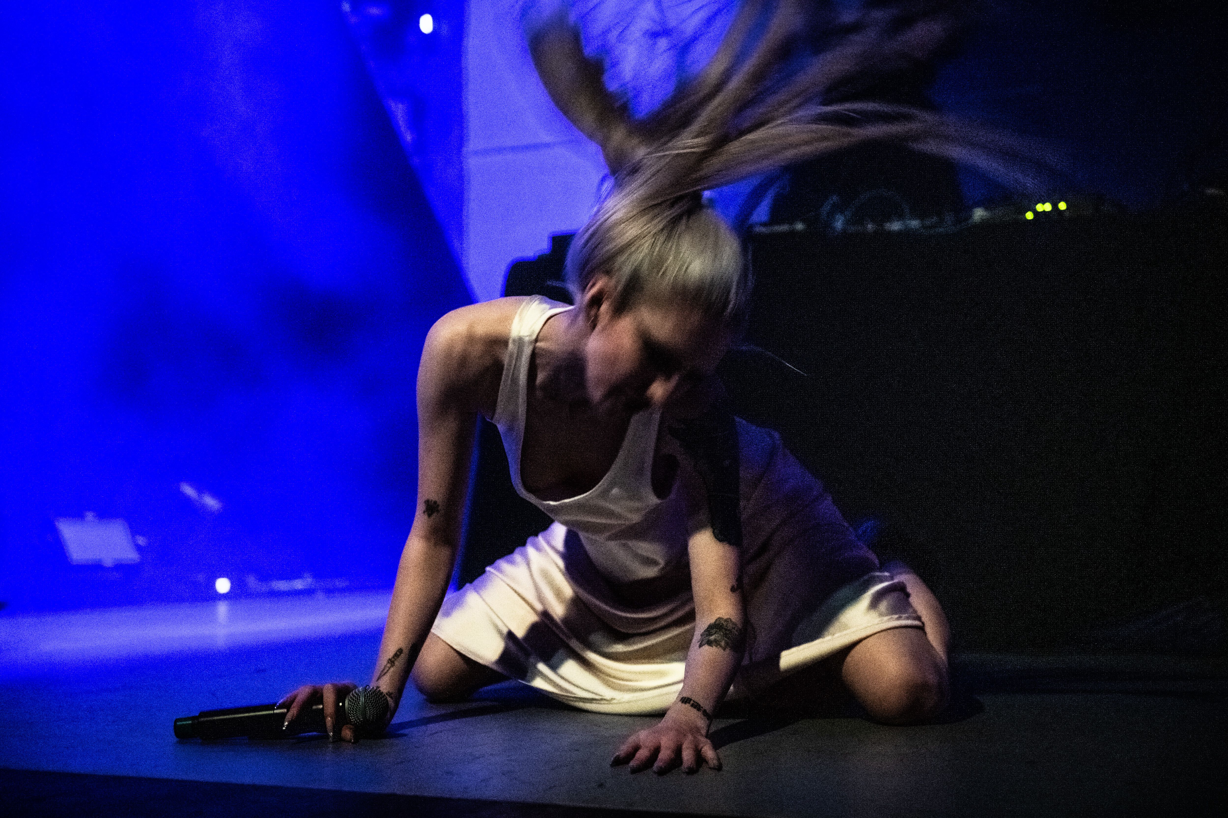 A person on a stage is captured mid-motion, their long hair flying upward as they crouch near a microphone. The scene is drenched in deep blue light, creating a high-energy, cinematic atmosphere.