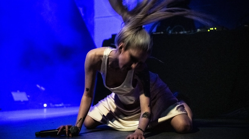 A person on a stage is captured mid-motion, their long hair flying upward as they crouch near a microphone. The scene is drenched in deep blue light, creating a high-energy, cinematic atmosphere.