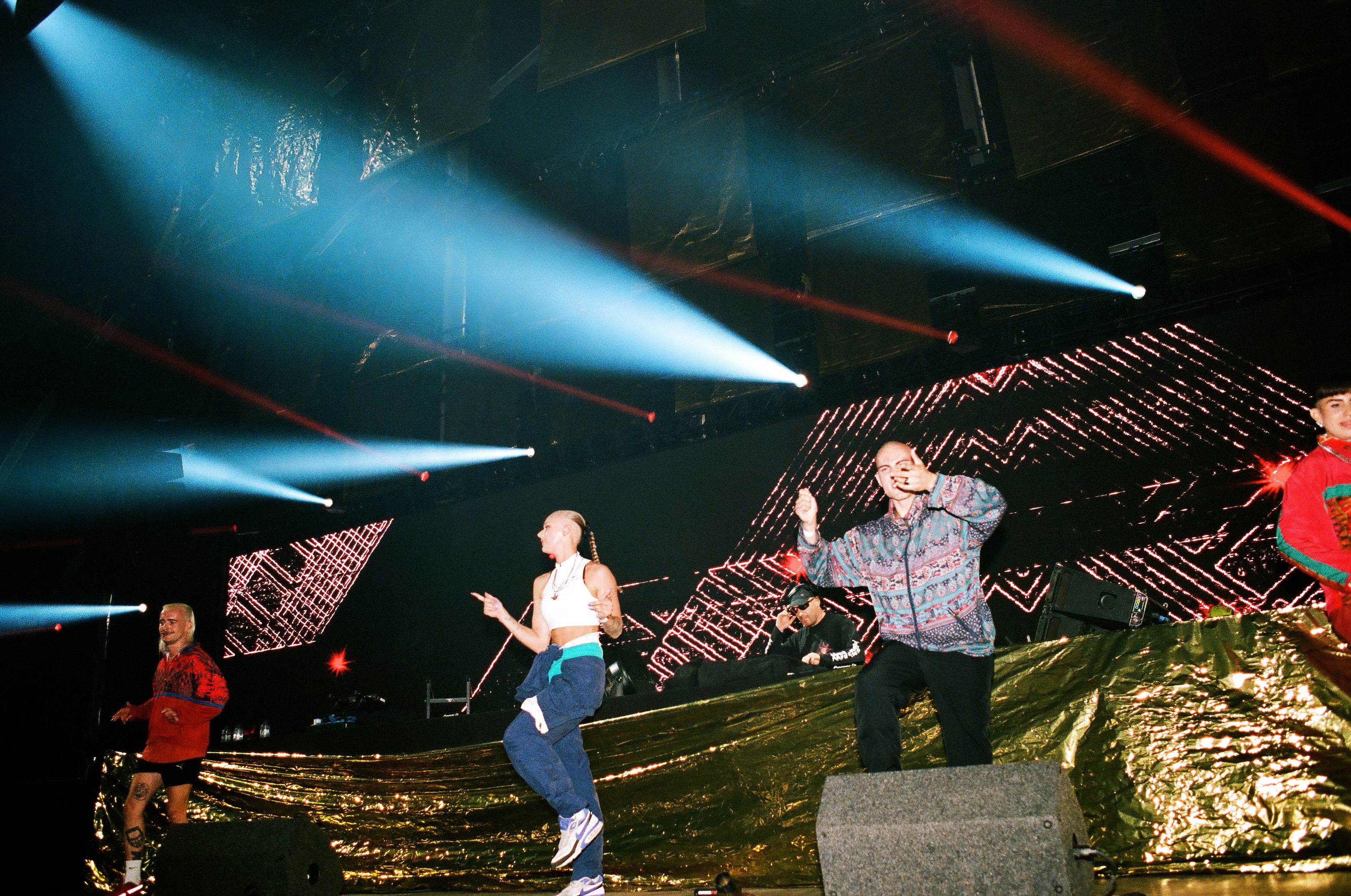 Dancers in colorful tracksuits perform on a stage with a gold-foil backdrop and large LED screens. Bright blue spotlights cut through the dark venue, creating a high-energy rave atmosphere.