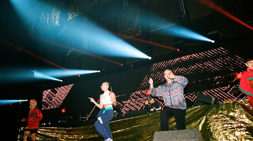 Dancers in colorful tracksuits perform on a stage with a gold-foil backdrop and large LED screens. Bright blue spotlights cut through the dark venue, creating a high-energy rave atmosphere.