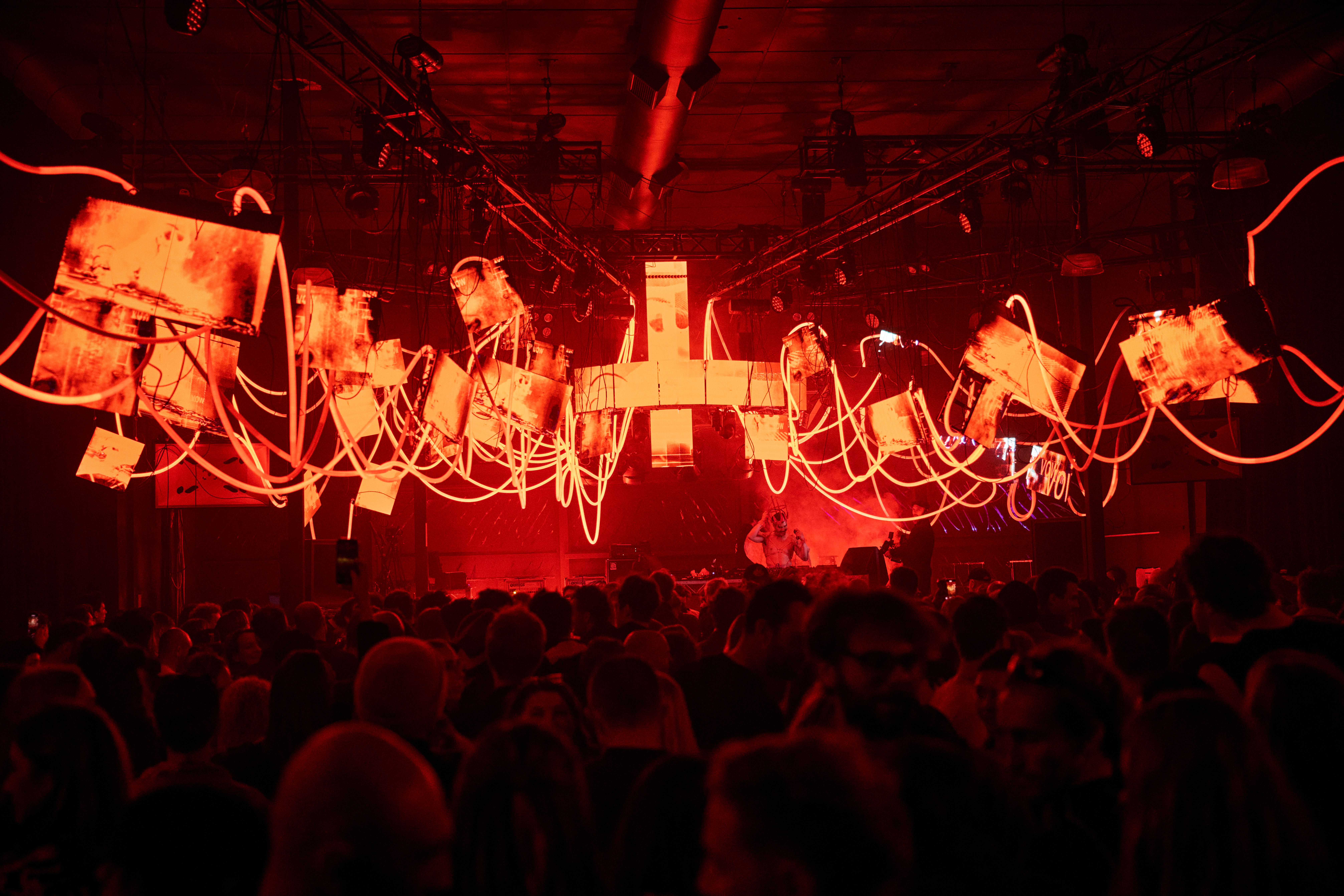A large crowd at Night Mass is bathed in deep red light, with glowing neon tubes and screens suspended haphazardly from the ceiling above.