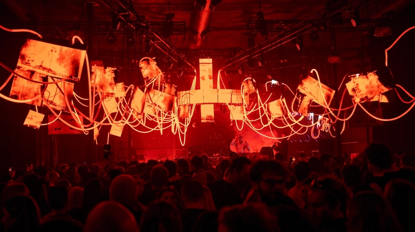 A large crowd at Night Mass is bathed in deep red light, with glowing neon tubes and screens suspended haphazardly from the ceiling above.