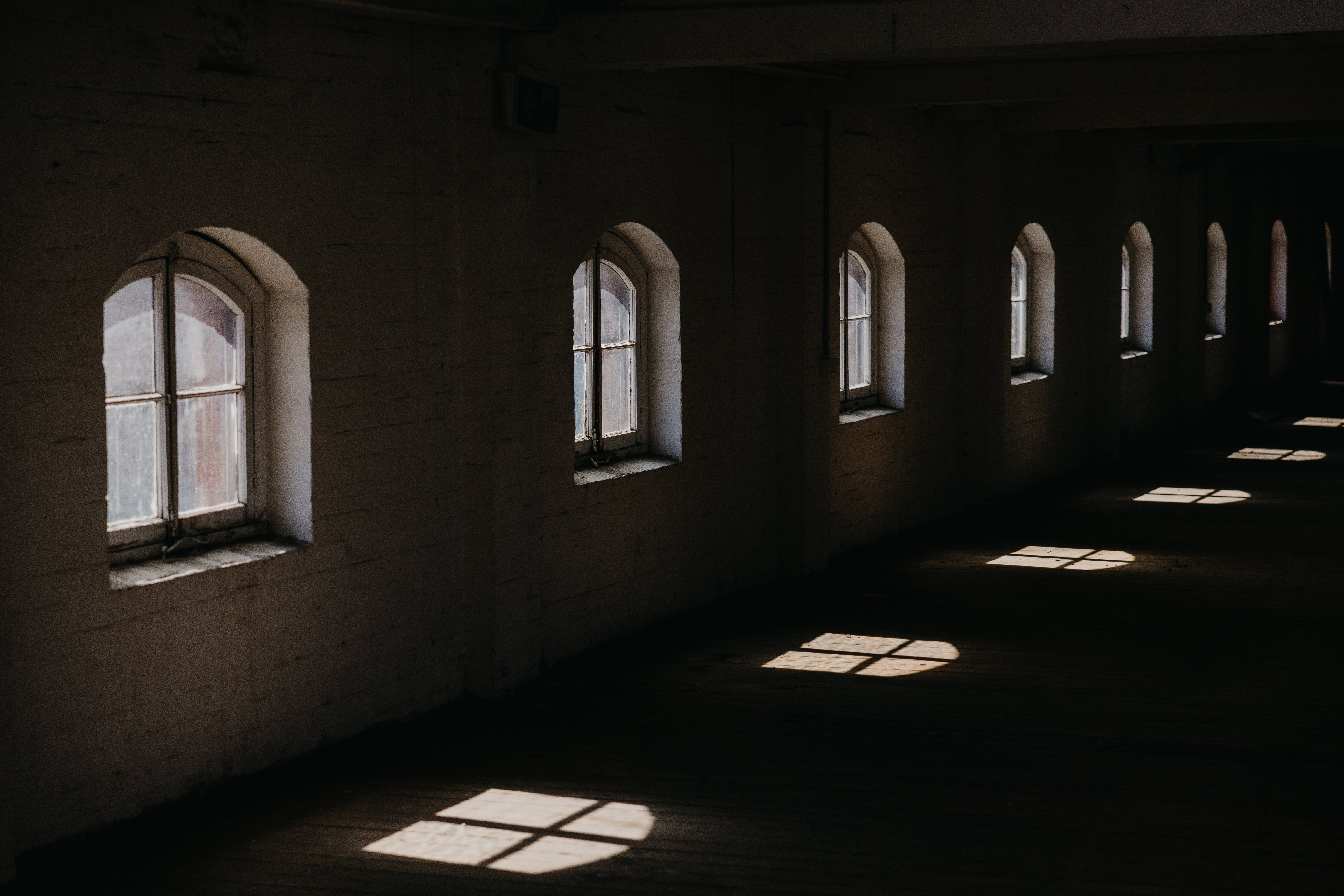 A row of arched windows in a dark, empty warehouse allows sunlight to cast bright, geometric patterns across the wooden floor.