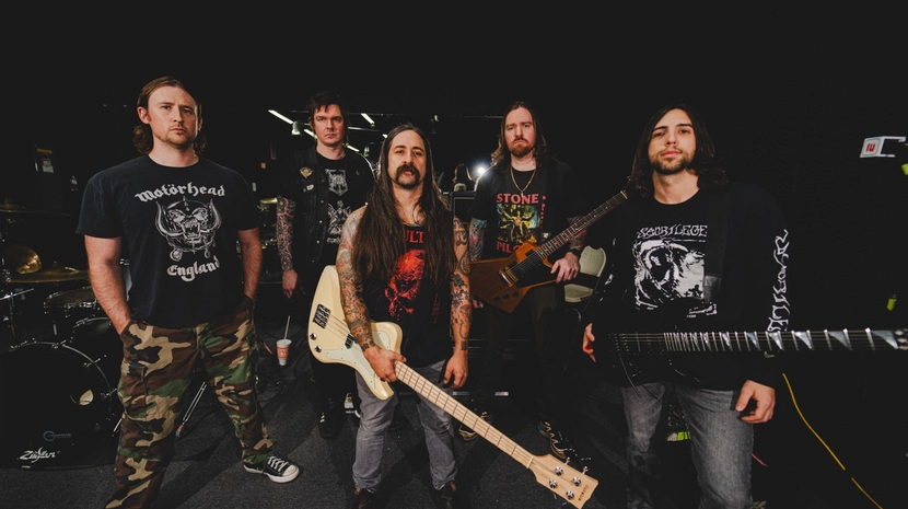 The five members of a metal band pose with their instruments in a dark studio, wearing band t-shirts and casual gear.