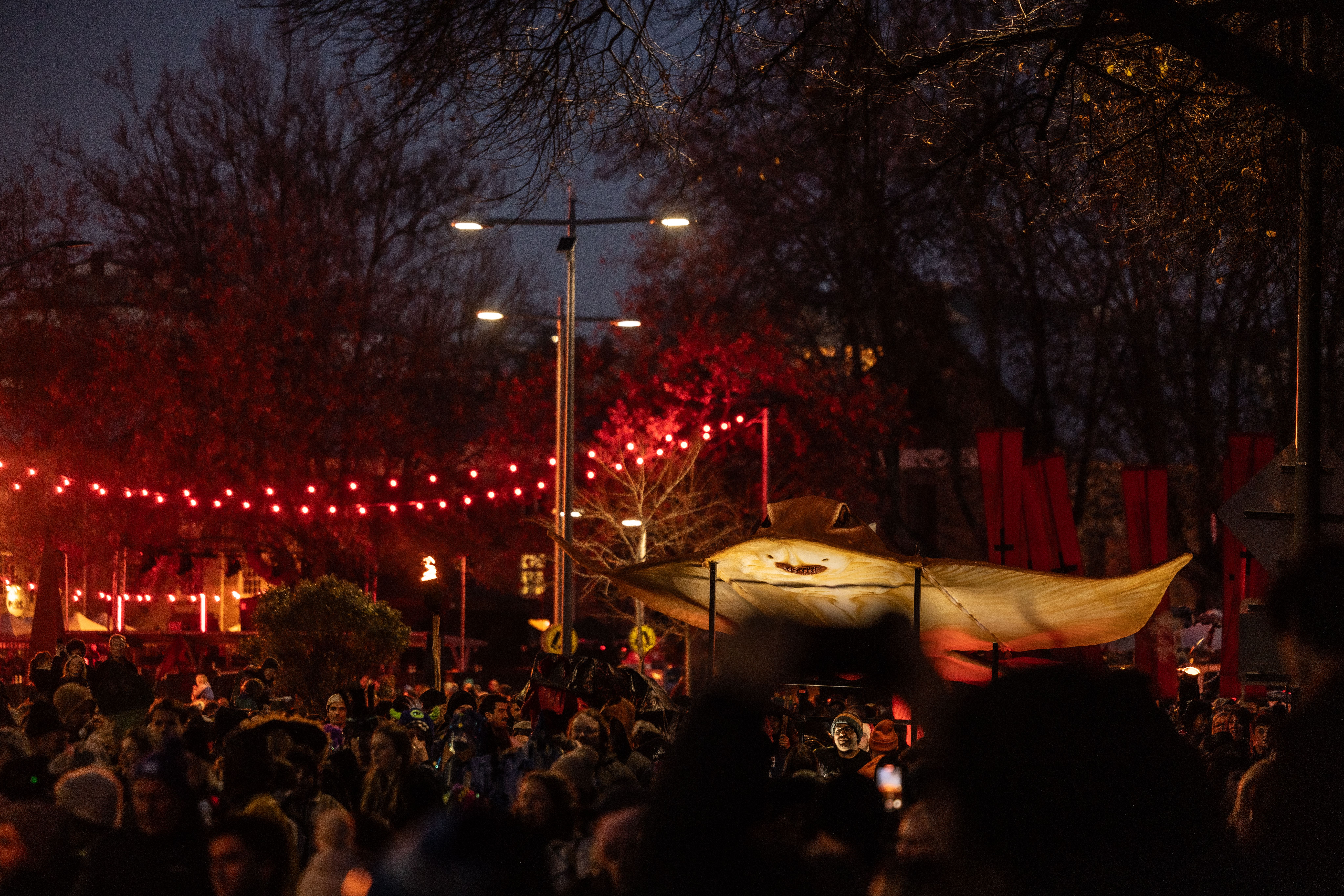 A large, illuminated skate ogoh is carried in a crowded outdoor procession, decorated with red string lights and dark trees.