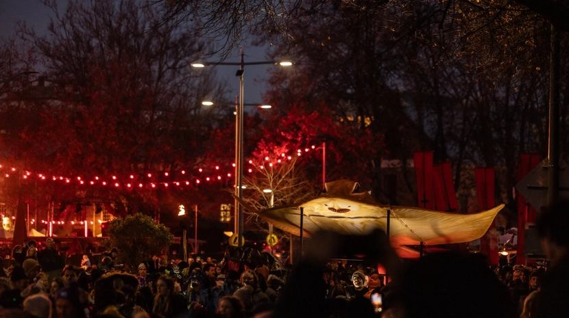 A large, illuminated skate ogoh is carried in a crowded outdoor procession, decorated with red string lights and dark trees.